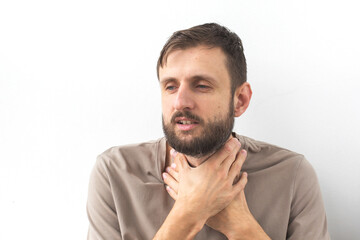 Naklejka premium Bearded man in a beige T-shirt holding his throat with both hands, looking unwell. His facial expression suggests discomfort or pain. Concept of health, illness, sore throat