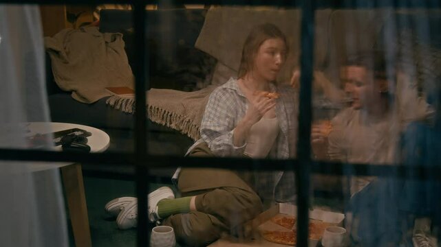 Full shot, watched outside through window on rainy night, of young Caucasian couple sitting on floor at home, enjoying takeaway pizza from box, having fun conversation while chewing