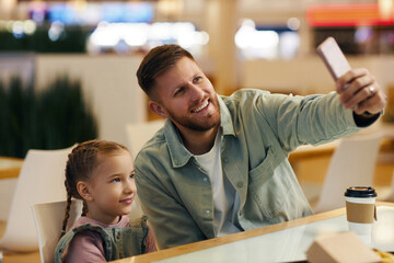 Medium close up of young man smiling widely while taking selfie with his beautiful daughter who sitting near him at food court
