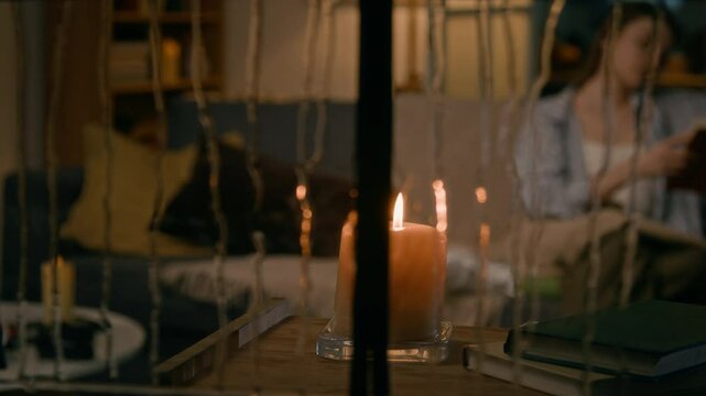 Full panning shot of candle burning on table by window on rainy night, and young woman sitting on couch with blanket, reading book in blurred background, seen by stranger or stalker through window