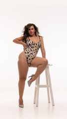 Plus size woman leaning on white stool, wearing animal print bodysuit and striped heels, showcasing...