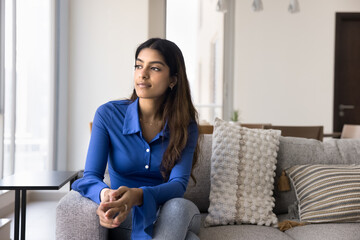 Young Indian woman dressed casually sit on couch in relaxed posture, gaze thoughtfully out of...