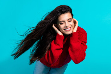 Stylish young woman in red sweater and jeans smiling happily against a turquoise background,...