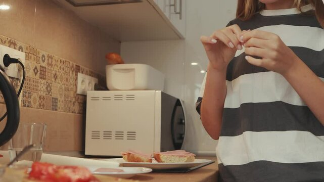 Teenage female preparing ham and cheese sandwich, spreading condiments in sleek kitchen, smiling while assembling fresh ingredients