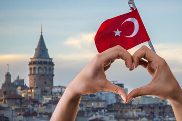 Heart shape hand gesture against the backdrop of Galata Tower and Turkish flag in Istanbul city view.