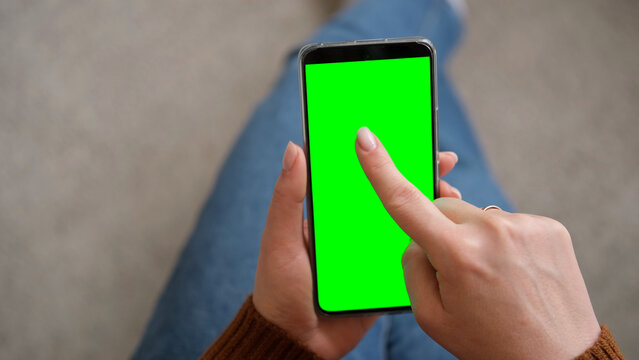 Young woman sitting at home holding smartphone green mock-up screen in hand. Female person use chroma key mobile phone. Vertical mode. Touching, swiping display, tapping, surfing internet social media