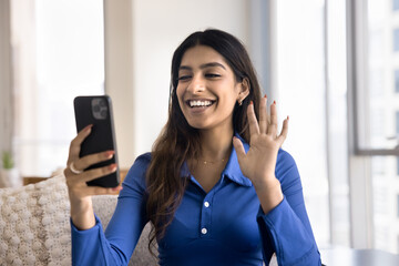 Pretty Indian female holding smartphone, smiling, greeting friend, start real-time remote videocall conversation seated on couch at home. Worldwide communication using modern wireless tech and apps