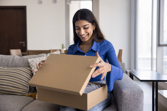 Young Indian woman opening cardboard box, received parcel with long-awaited clothes or bought items for house, smile enjoy moment of unpacking, feel satisfied with quality and express delivery service