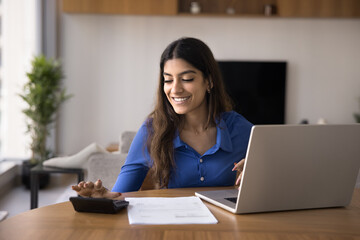 Indian woman sits at desk with laptop, using calculator, calculating expenses, organizing or...
