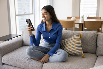 Obraz premium Attractive young 20s Indian woman using smartphone seated on cozy sofa in modern living room, smile, check messages, browsing social media, watch videos, reading latest on-line news on weekend at home