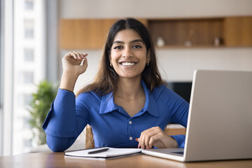 Portrait of motivated young Indian woman sit at desk with laptop and notebook in front of her,...