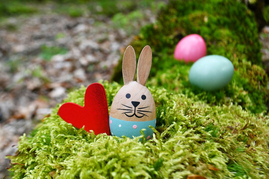 Kleiner Osterhase mit rotem Herz aus Holz, zwei gef&auml;rbte Eier im Hintergrund, im Wald