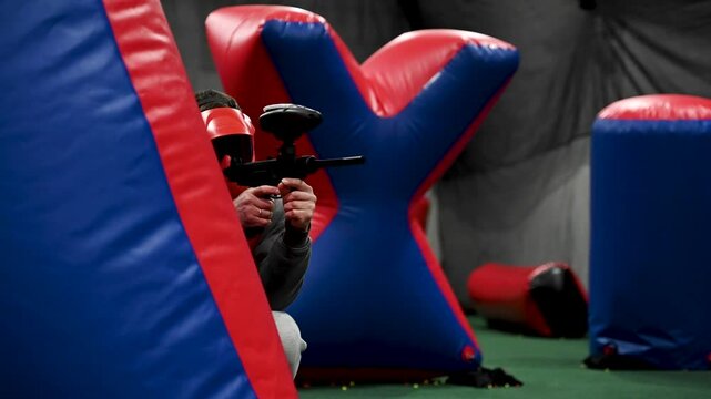 A paintball player wearing a mask and carrying a gun hides behind soft fences from other paintball players to avoid being shot.
