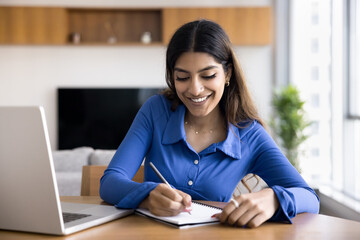 Woman smiles while writes in notebook seated at desk with laptop, takes notes, make plan,...