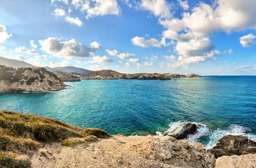 Breathtaking coastal landscape with turquoise sea, rugged cliffs, and a distant town under a bright blue sky with fluffy clouds. Waves crash against rocks, creating a serene and picturesque view.