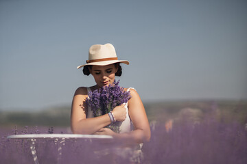 woman sitting in a field of lavender and wearing a straw hat. She is smiling and holding a bouquet of flowers. Scene is peaceful and serene, as the woman is surrounded by the beauty of nature