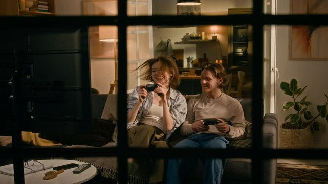 Full shot, seen through window, of young Caucasian couple sitting together on couch at home in evening night, playing video game together on TV, girl winning, cheering and whooping with joy