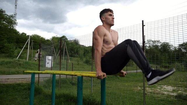 Strong guy training abdominal muscles on parallel bars at sports ground. Young shirtless sportsman exercising at nature. Sporty athlete working out at nature. Concept of active sportive lifestyle