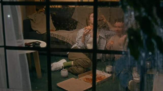 Full shot, seen outside through window, of young Caucasian woman and man sitting on floor in living room at home, during rainy evening, eating takeaway pizza from box, chatting and laughing
