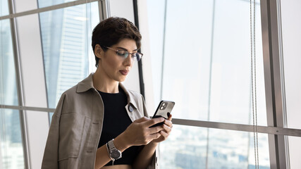 Business networking on the go. Mature businesswoman entrepreneur use phone by window wall in airport to stay connected check flight schedule pay for service book tickets via text message. Copy space