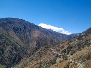  A winding dirt road traverses the rugged Andean mountains under a clear blue sky. The dry, rocky terrain contrasts with patches of greenery on steep slopes