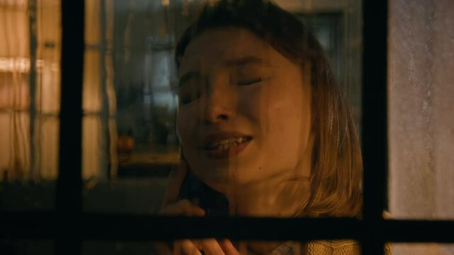 Closeup of young Caucasian woman making phone call while standing by window in rainy night, arguing with husband, then frantically screaming, shouting in anger and crying desperately