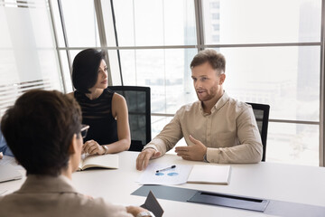 Diverse team collaboration. Confident millennial man entrepreneur manager expert talk present creative business idea project solution to coworkers colleagues group gathered at table in conference room