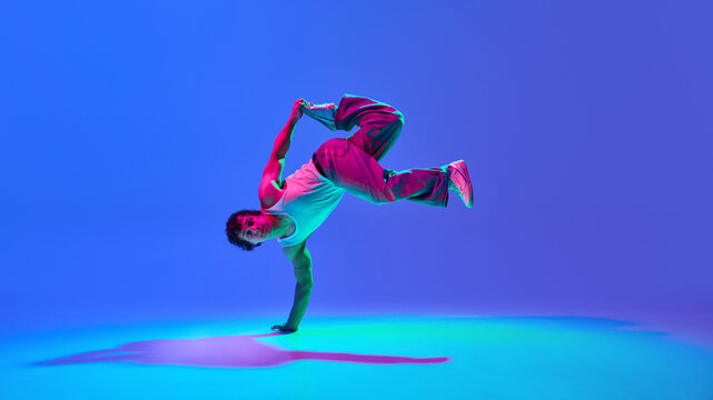 Burst of energy. Talented young guy, contemp dancer in motion, performing against gradient blue purple background in neon light. Concept of dance, street style, creativity, youth culture, energy