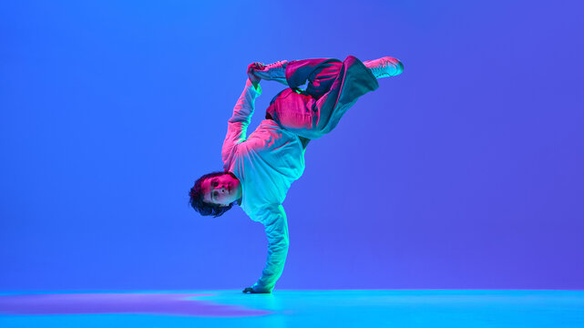 Young man, breakdancer blending flexibility, power, and rhythm, performing against gradient blue purple background in neon light. Concept of dance, street style, creativity, youth culture, energy