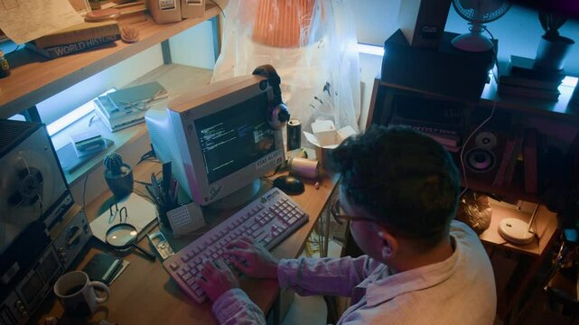 High angle view of young male IT specialist writing code in obsolete programming language on retro computer while sitting at wooden desk in small room