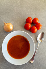 A bowl of freshly made tomato soup is placed on a grey countertop, accompanied by a whole onion and ripe red tomatoes. 