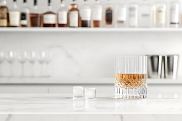 A crystal glass filled with amber liquor and two ice cubes sitting atop a modern white marble countertop bar top creates a simple and crisp upscale atmosphere.