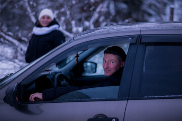 A man peeks out of his car on a snowy winter day, with a woman in the background.