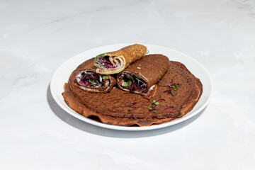 A plate of dark buckwheat or chocolate crepes, neatly arranged and garnished with fresh microgreens, featuring a healthy vegetable filling.
