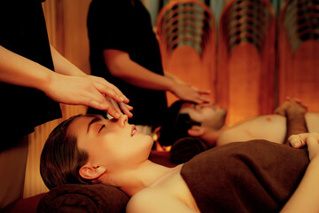 Couple customer enjoying relaxing anti-stress head massage and pampering facial beauty skin recreation leisure in warm candle lighting ambient salon spa in luxury resort or hotel. Quiescent