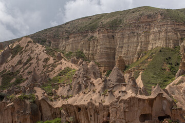 Ancient rock formations with carved dwellings rise over a green valley. A distant town sits at the foot of rolling hills under a cloudy sky, blending history, nature, and human presence in a breathtak