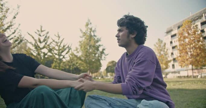 A young couple sits in the park, holding hands and talking. They share a kiss and continue their conversation.