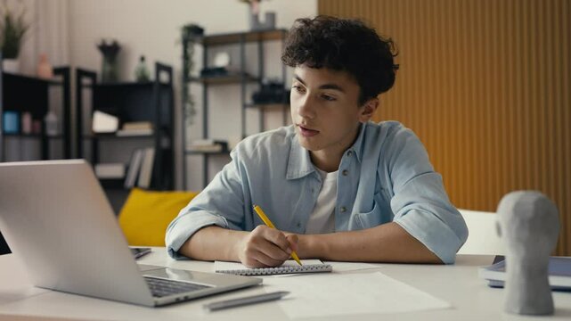 Teenager studying at home, writing in a notebook and using a laptop for research