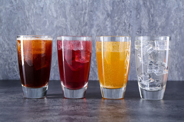Sweet soda water of different flavors with ice cubes in glasses on grey table