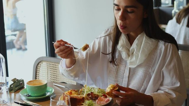 A woman savoring a cheesy brunch in a stylish cafe. She takes a bite from her dish while sipping coffee, immersed in her meal and surroundings.