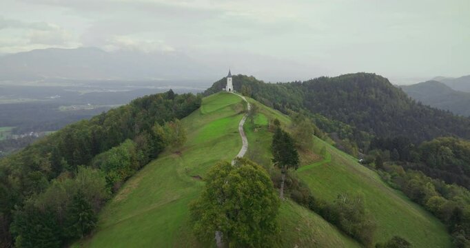 The Church of Saint Primus and Felician in Jamnik, Slovenia, is isolated among lush green hills. The impressive view is revealed as the drone flies along the winding path towards the church.