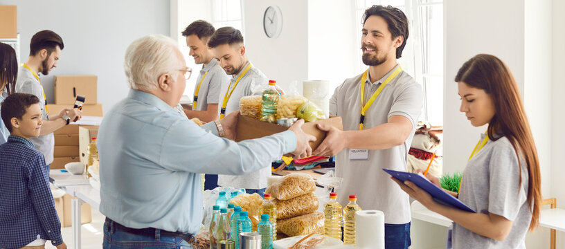 Young cheerful man working at charity center with a team of volunteers and giving free food donations in charitable foundation for people in need. Humanitarian aid and volunteering concept. Banner.
