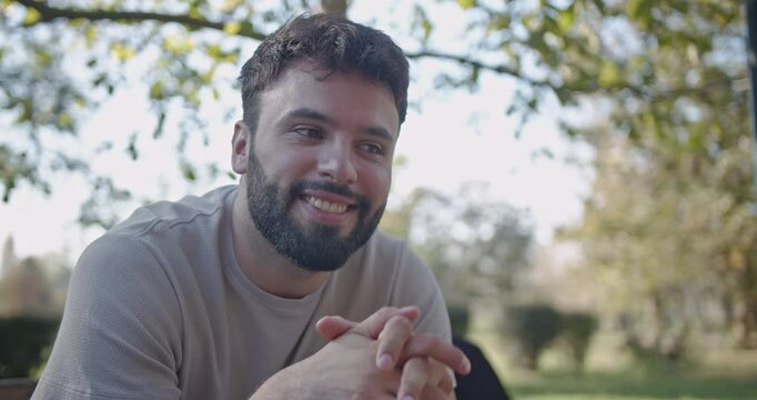A young man sits on a bench in the park and expresses different emotions, going from happiness to sadness, then thoughtful.