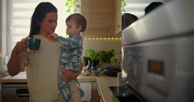 Mother holding baby in kitchen as child reaches for hot cup, protective parenting moment during everyday family routine, nurturing and safety in a warm home setting