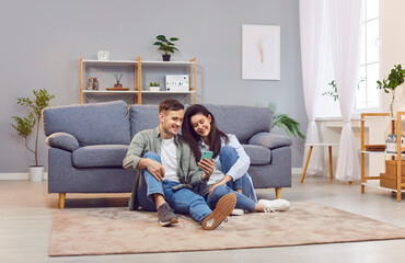 Happy couple at home, young woman and man floor sitting close in new modern living room, using...