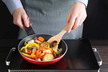 Woman frying vegetables and mushrooms in pan on stove at wooden table, closeup