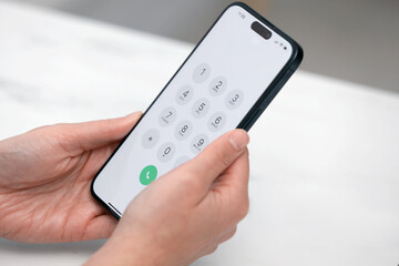 Woman holding smartphone with dialer application at white table against blurred background, closeup