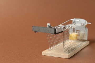 Metal mouse trap with piece of cheese on brown background, closeup. Space for text