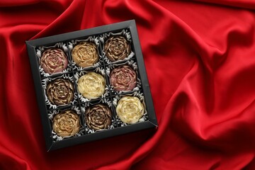 Delicious flower shaped chocolate bonbons in box on red fabric background, top view. Space for text