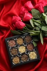 Delicious flower shaped chocolate bonbons in box and roses on red fabric background, top view